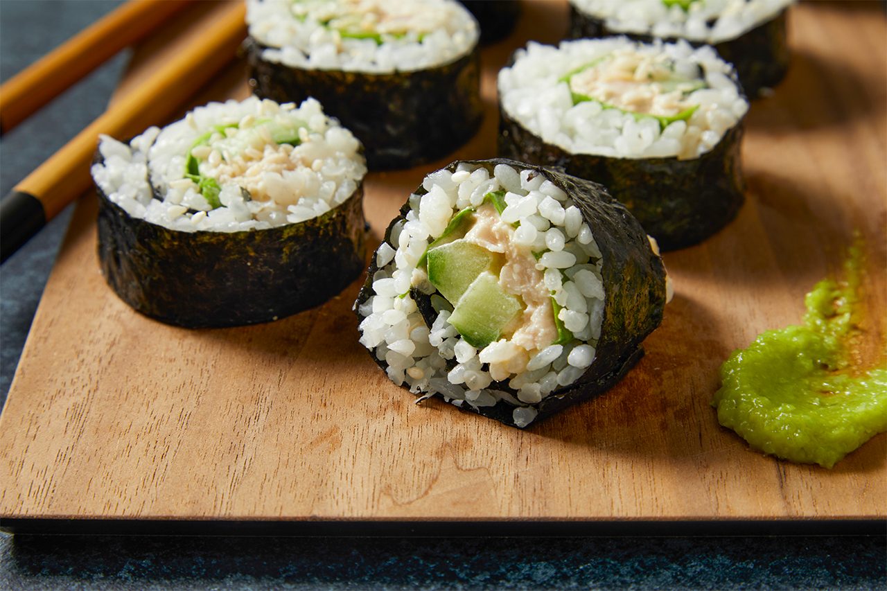 Eye Level horizontal image of several sushi rolls presented on a wooden cutting board. The maki rolls feature white rice, nori, diced cucumber, and a creamy, shredded filling, possibly chicken or tuna. A small dollop of bright green wasabi is visible beside one roll, with chopsticks resting in the background.