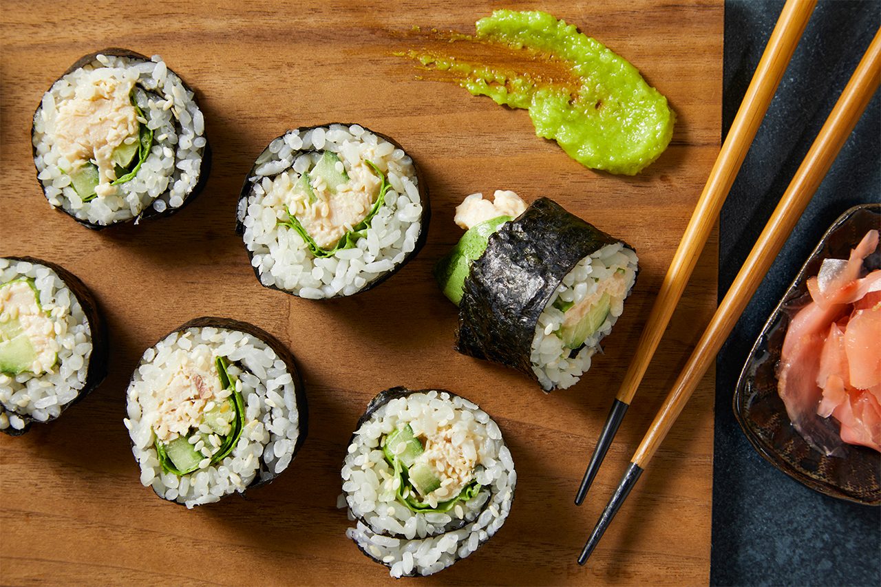 Overhead horizontal image of several maki-style sushi rolls on a wooden surface. The rolls feature rice, nori, a creamy filling, cucumber, and lettuce, garnished with sesame seeds. A smear of wasabi, a pair of chopsticks, and a small dish of pickled ginger accompany the sushi.