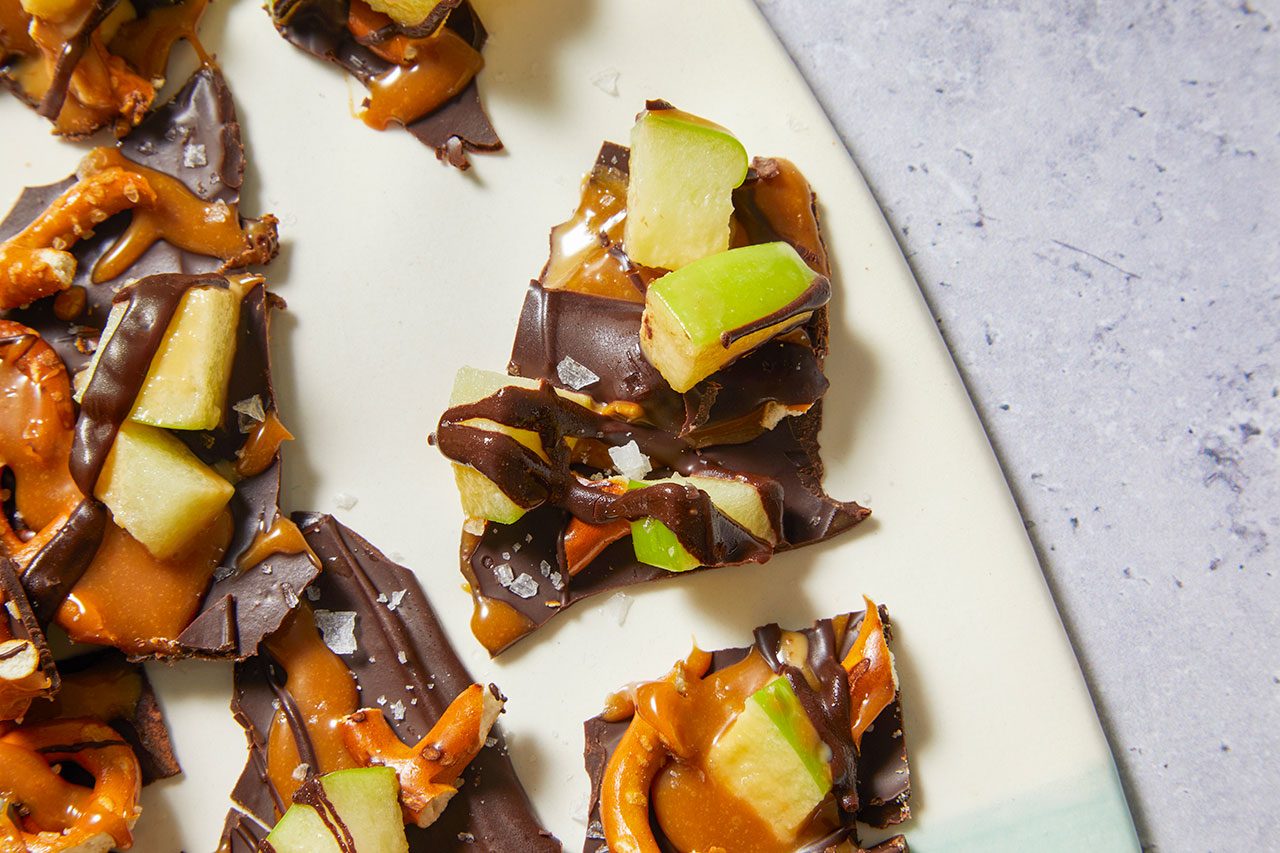 Overhead horizontal image of broken pieces of chocolate bark arranged on a white serving platter. The dark chocolate bark is topped with gooey caramel, broken pretzel pieces, diced green apple, a drizzle of dark chocolate, and sprinkled with sea salt.