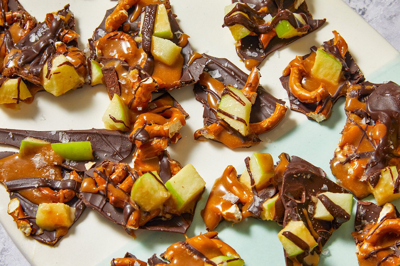 Overhead horizontal image of several pieces of chocolate bark. The dark chocolate bark is topped with golden caramel, crunchy pretzel pieces, diced green apples, and sprinkled with flaky sea salt.