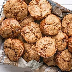 A basket filled with several golden-brown muffins, some in paper liners, displayed on a plaid cloth. The muffins have a crumbly, textured top and are arranged closely together.