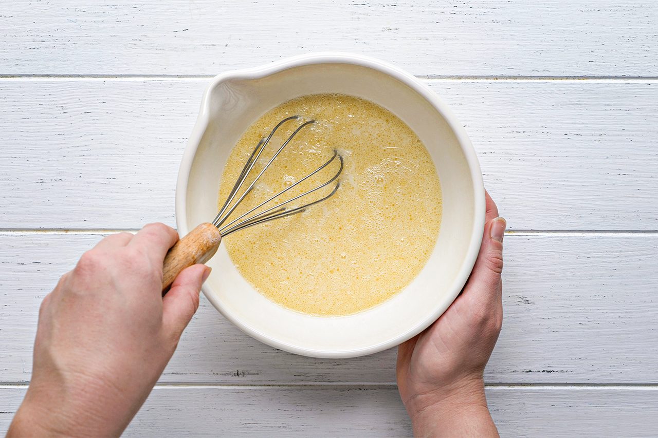 A person holds a white bowl and uses a whisk to mix a yellow batter on a white wooden surface.