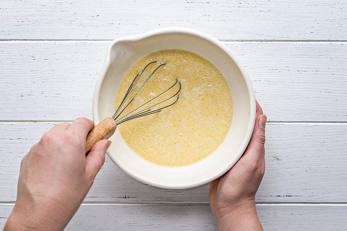 A person holds a white bowl and uses a whisk to mix a yellow batter on a white wooden surface.