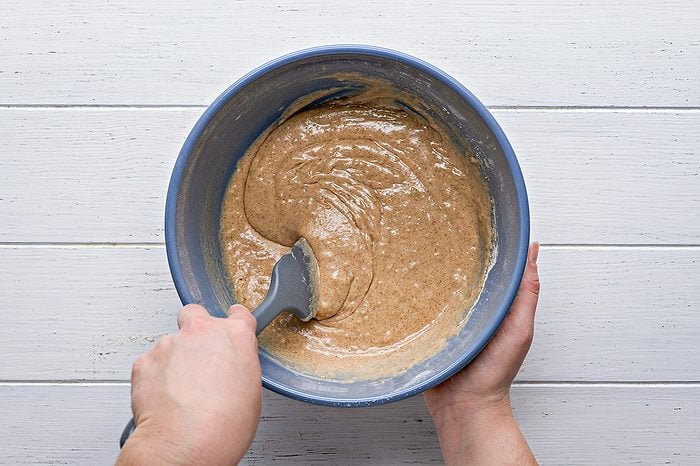 A person mixing light brown batter in a blue bowl with a spatula on a white wooden surface.