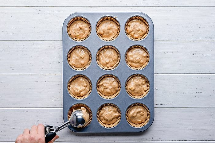 A hand uses an ice cream scoop to fill a muffin tin lined with paper cups and filled with muffin batter, on a white wooden surface.