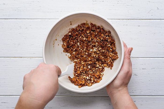 A person mixing granola ingredients in a white bowl with a white spatula on a white wooden surface.