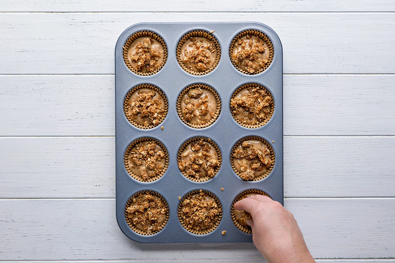 A hand reaches for one of twelve muffins in a muffin tin on a white wooden surface. The muffins have a crumbly topping.
