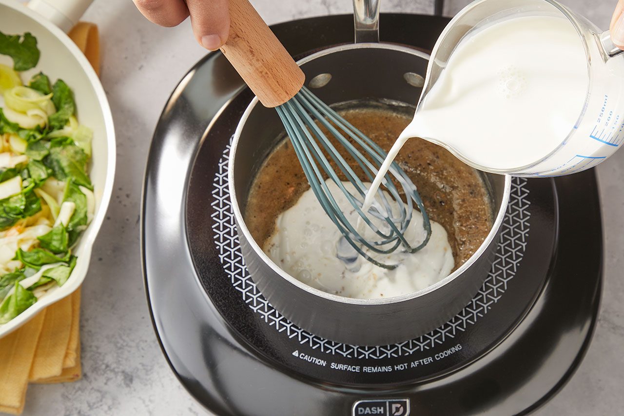 Overhead horizontal image of a hand pouring milk from a glass measuring cup into a saucepan. A whisk is used to combine the milk with a brown sauce base in the pot. In the background, a white bowl contains cooked green vegetables like bok choy and spinach.