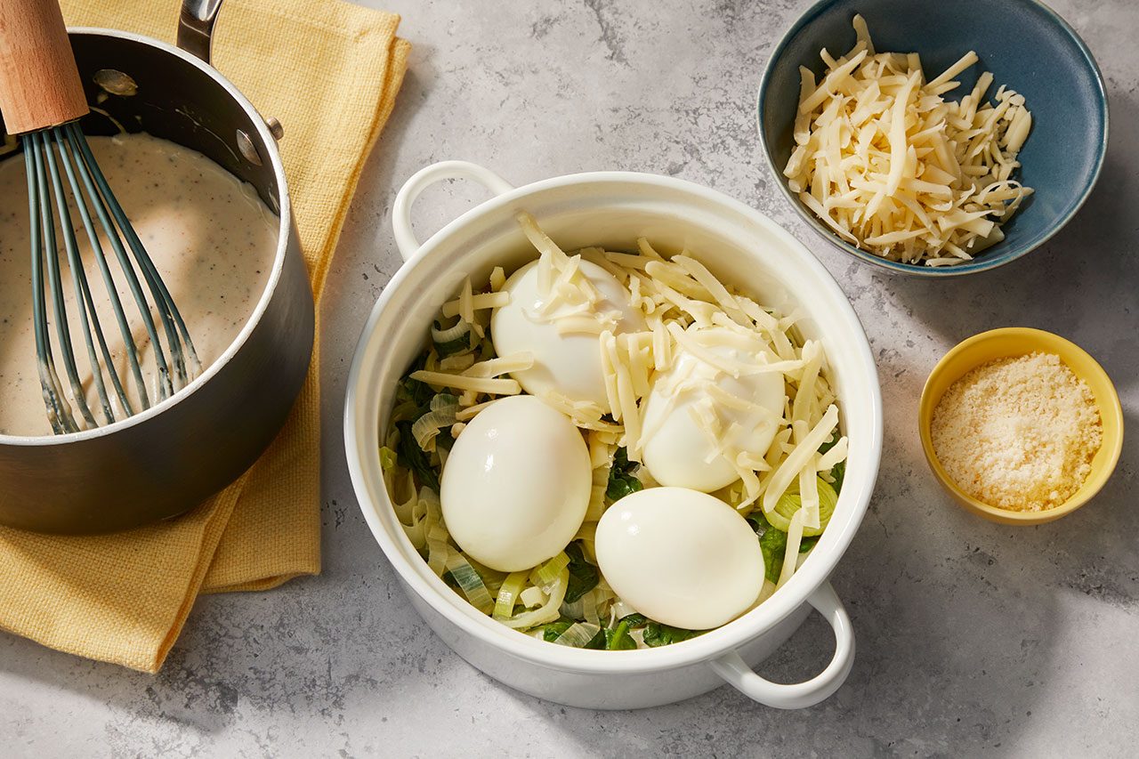 Overhead horizontal image of a creamy sauce in a saucepan with a whisk, positioned next to a white baking dish. The baking dish contains peeled hard-boiled eggs over chopped leeks, topped with shredded cheese. Additional bowls hold more shredded cheese and grated Parmesan cheese.
