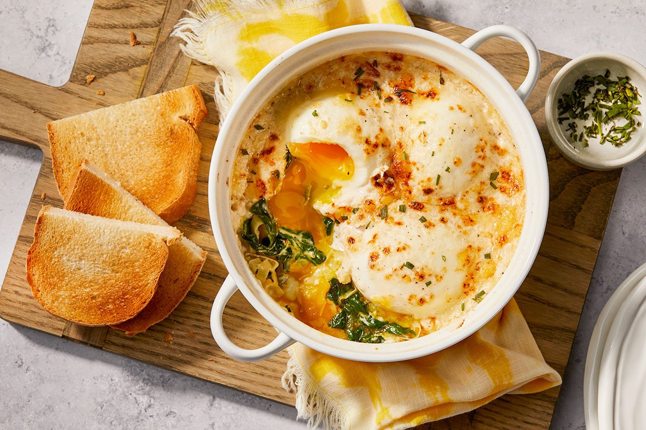 Overhead horizontal image of baked eggs with a runny yolk, spinach, and melted cheese in a white ramekin. It is served with toasted bread triangles on a wooden board, accompanied by a small bowl of dried herbs.