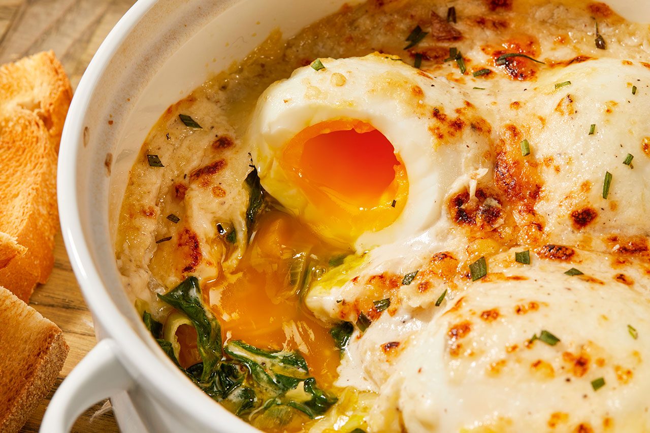 Overhead horizontal image of baked eggs in a white ramekin. The dish features melted, golden-brown cheese, and a broken egg reveals a rich, runny yolk amidst cooked greens and a creamy sauce, with golden-brown toasted bread on the side.