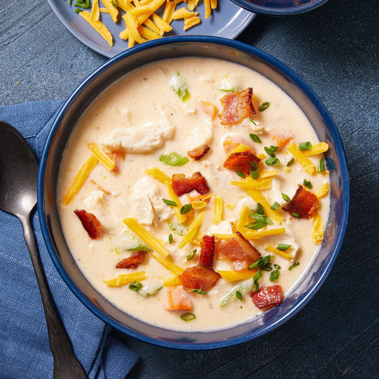 Overhead vertical image of a hearty bowl of creamy soup, garnished generously with shredded cheese, crispy bacon pieces, and chopped green onions. On the side, small dishes hold extra shredded cheese and crispy bacon, alongside a dark spoon on a blue napkin.