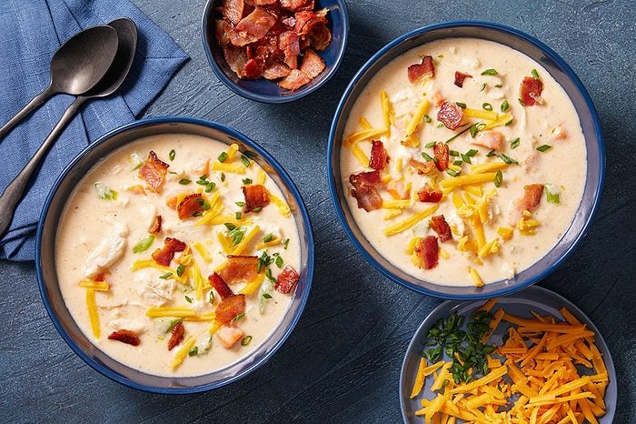 Overhead horizontal image of two bowls of creamy soup, garnished with shredded cheddar cheese, crumbled bacon, and chopped chives. A small bowl of crumbled bacon and a plate with more shredded cheese and chives are visible, alongside two dark spoons on a blue napkin.