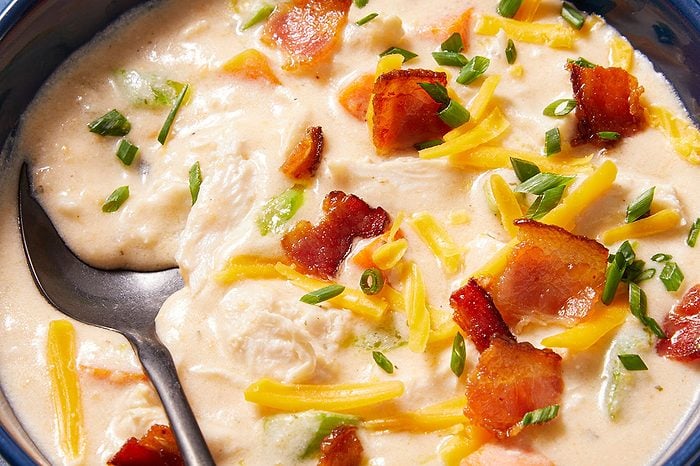Overhead horizontal image of a rich, creamy soup in a dark blue bowl, garnished generously with crispy bacon bits, shredded yellow cheese, and chopped green onions. A black spoon rests in the left side of the bowl, within the hearty soup that also contains visible pieces of chicken and potato.