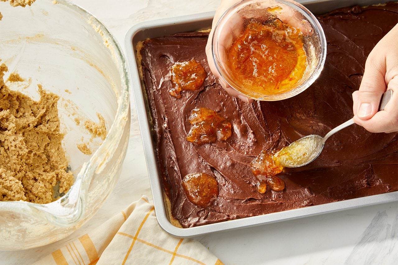 Overhead horizontal image of a hand spooning bright orange marmalade from a glass bowl onto a rich chocolate layer in a rectangular baking pan. Several dollops of the vibrant jam are already placed on the dark chocolate, next to a bowl of cookie dough and a yellow kitchen towel. | [Original] RMS 283594 Chocolate Orange Oat Bars; How to; Step 4 Spread over crust. Dollop with orange marmalade. Sprinkle with remaining oat mixture