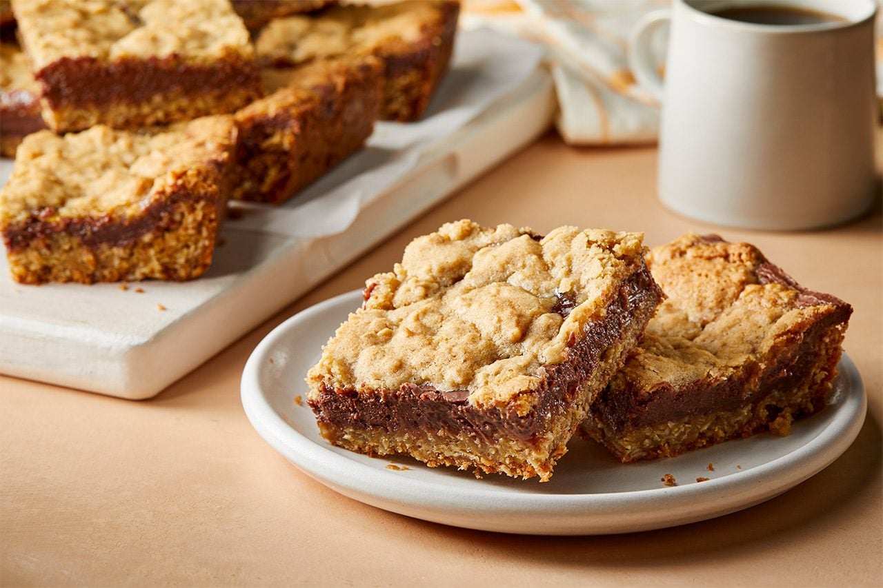 Eye level horizontal image of two square dessert bars with an oatmeal cookie base and a rich chocolate filling layer, presented on a small white plate. In the background, more bars are visible on a white cutting board lined with parchment paper, alongside a white mug filled with coffee. | [Original] RMS 283594 Chocolate Orange Oat Bars; Horizontal AP; Pull