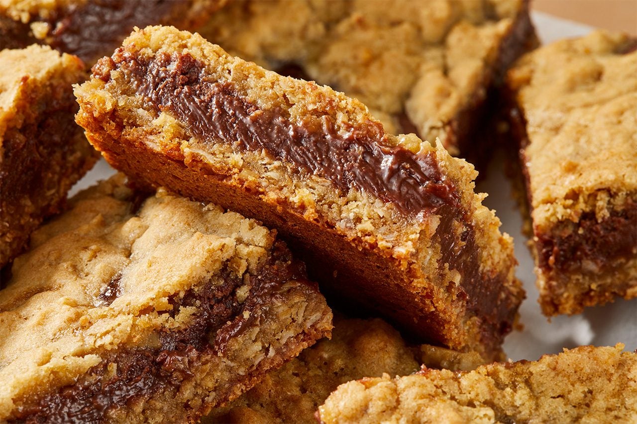 Eye Level horizontal image of several chocolate-filled dessert bars. One bar is prominently propped up, showcasing its thick, rich chocolate filling sandwiched between two layers of golden-brown crust with visible texture, likely containing oats and coconut. | [Original] RMS 283594 Chocolate Orange Oat Bars; Horizontal; PUSH