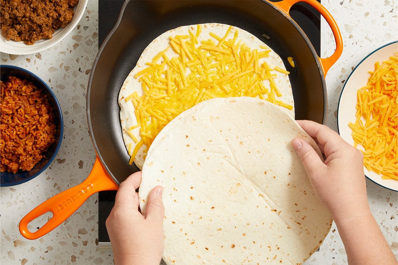 Overhead horizontal image of hands placing a flour tortilla over another flour tortilla filled with shredded cheddar cheese in an orange Le Creuset skillet. Bowls of cooked ground beef, Spanish rice, and additional shredded cheddar cheese are arranged around the pan on a speckled countertop.