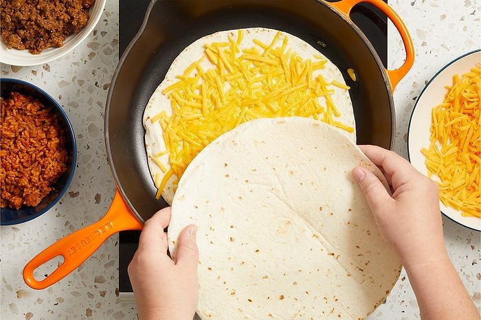 Overhead horizontal image of hands placing a flour tortilla over another flour tortilla filled with shredded cheddar cheese in an orange Le Creuset skillet. Bowls of cooked ground beef, Spanish rice, and additional shredded cheddar cheese are arranged around the pan on a speckled countertop.