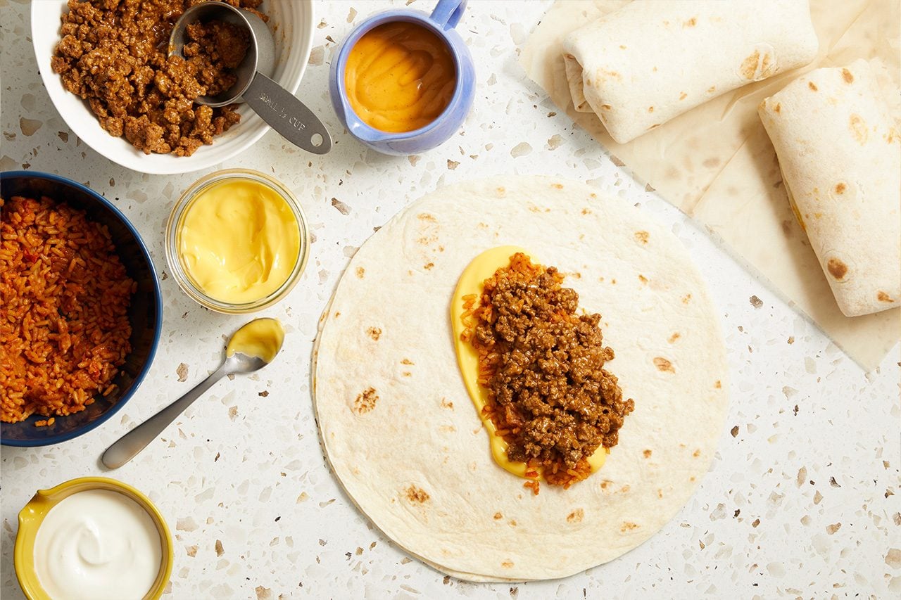 Overhead horizontal image of a flour tortilla being prepared, with ground taco meat and yellow cheese sauce spooned onto it. Bowls containing ground taco meat, Spanish rice, additional yellow cheese sauce, and sour cream are arranged around the tortilla, with two finished wrapped burritos visible on the right.