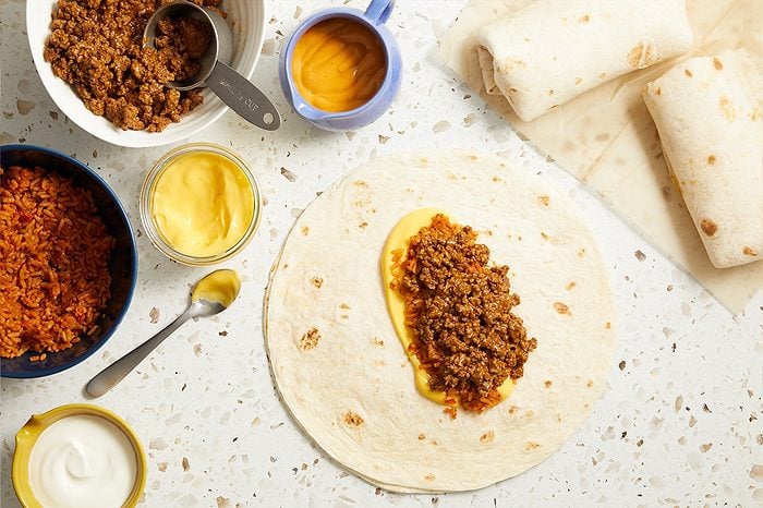 Overhead horizontal image of a flour tortilla being prepared, with ground taco meat and yellow cheese sauce spooned onto it. Bowls containing ground taco meat, Spanish rice, additional yellow cheese sauce, and sour cream are arranged around the tortilla, with two finished wrapped burritos visible on the right.