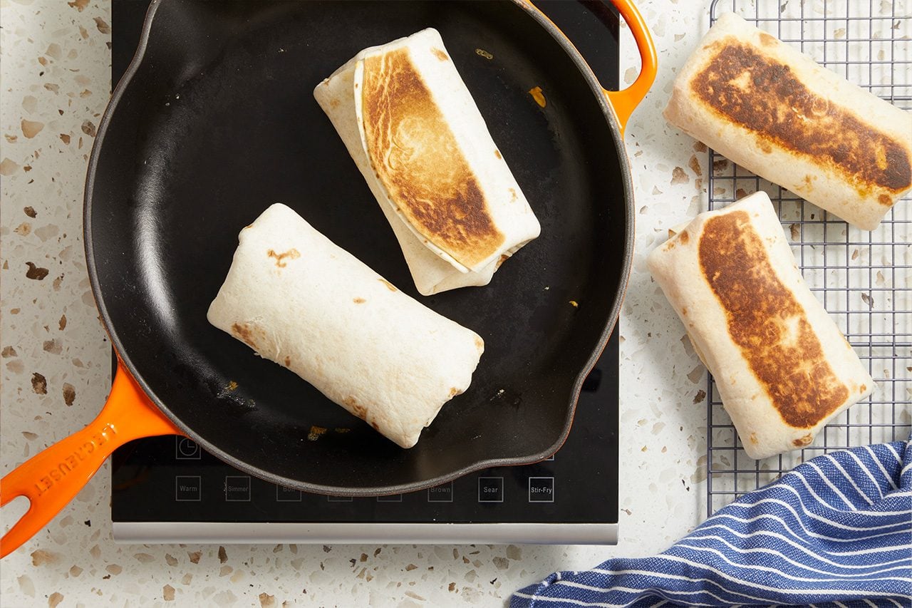 Overhead horizontal image of several burritos being toasted. Two burritos are browning in a black Le Creuset cast iron skillet on an induction cooktop, while two more burritos, already toasted, rest on a wire cooling rack next to a blue and white striped towel.