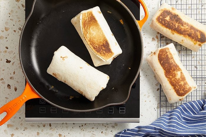 Overhead horizontal image of several burritos being toasted. Two burritos are browning in a black Le Creuset cast iron skillet on an induction cooktop, while two more burritos, already toasted, rest on a wire cooling rack next to a blue and white striped towel.