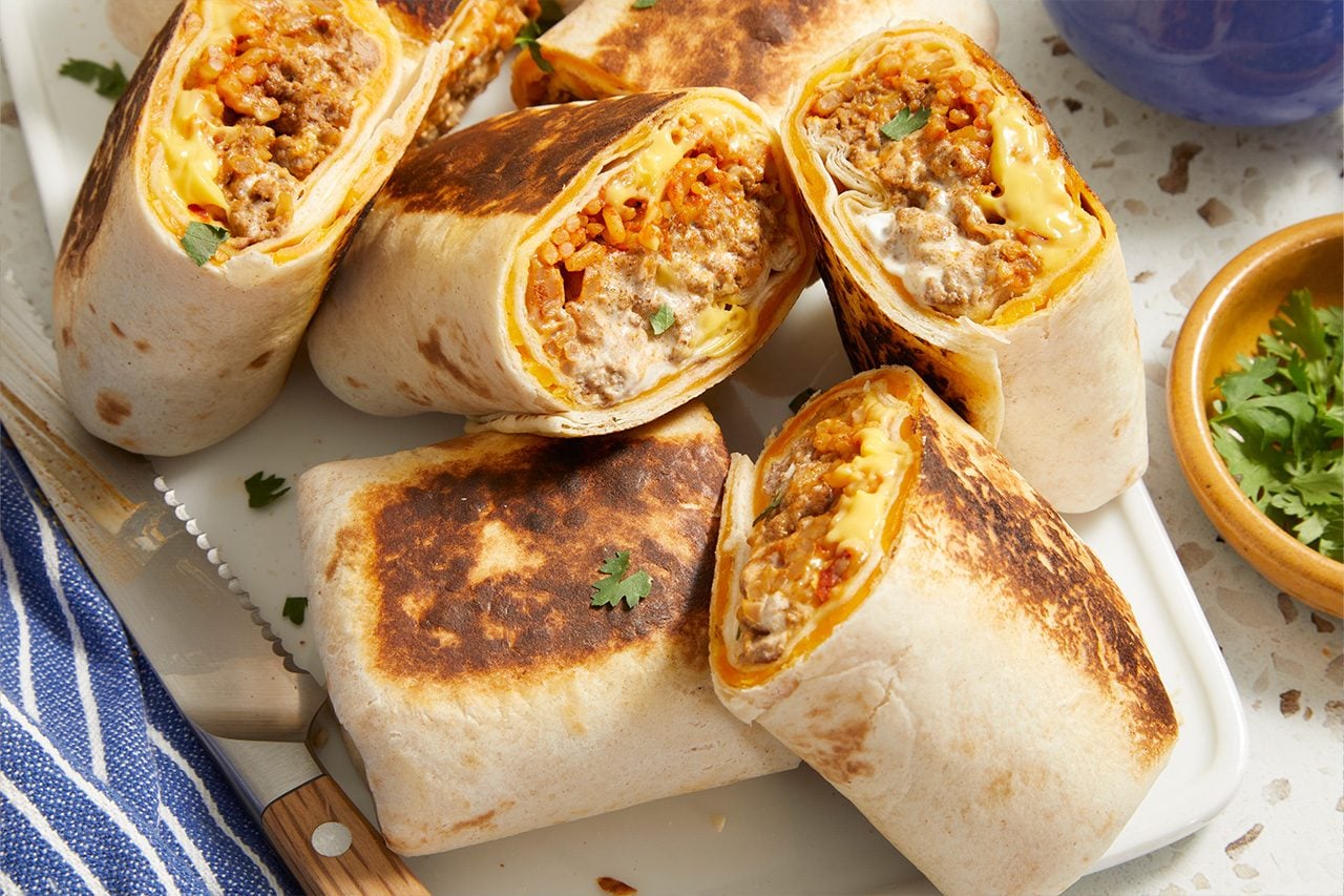 Eye-level horizontal image of several toasted burritos arranged on a white rectangular dish. Some of the burritos are cut in half, revealing a savory filling of ground meat, orange rice, melted cheese, and a creamy white sauce. Fresh cilantro sprigs garnish the dish and are also present in a small wooden bowl.