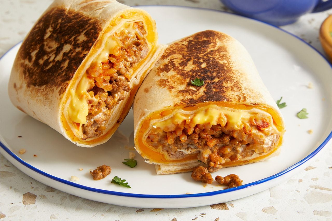 Eye level horizontal image of a large, toasted burrito cut in half, showcasing its savory interior. The filling is a hearty mix of seasoned ground meat, rice with a reddish sauce, and melted yellow cheese, all wrapped in a golden-brown tortilla. A few sprigs of fresh parsley garnish the white plate with a blue rim.
