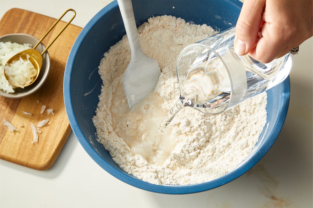 Overhead horizontal image of a hand pouring water from a measuring cup into a blue mixing bowl filled with flour. A white spatula is visible in the bowl, and a small bowl of diced onions sits on a wooden cutting board to the left. | [Original] RMS 283003 East African Rolex; Rolled Eggs; Contest-Everything Eggs; How to; Step 1 Make chapati dough - actionable shot is stirring water/onion OR kneading dough; Horizontal