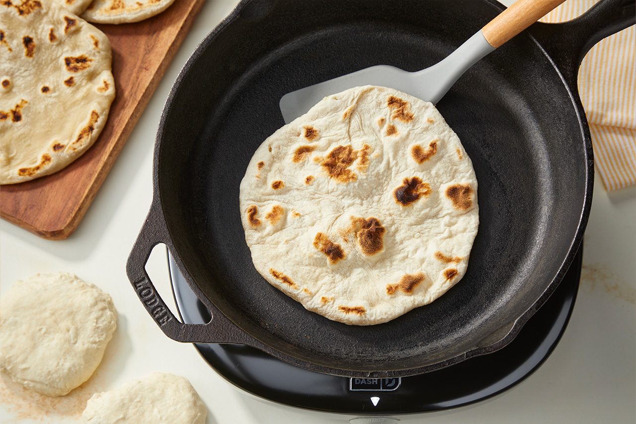 Overhead horizontal image of a cooked flatbread in a black cast iron skillet, with a gray spatula visible underneath it. Several golden-brown cooked flatbreads are on a wooden cutting board, and portions of raw dough are visible on the white counter. | [Original] RMS 283003 East African Rolex; Rolled Eggs; Contest-Everything Eggs; How to; Step 2 Portion and roll out into 6" rounds; Edge in some rolled and finished cooking; mainly show 1 or 2 being cooked in cast-iron pan; Horizontal
