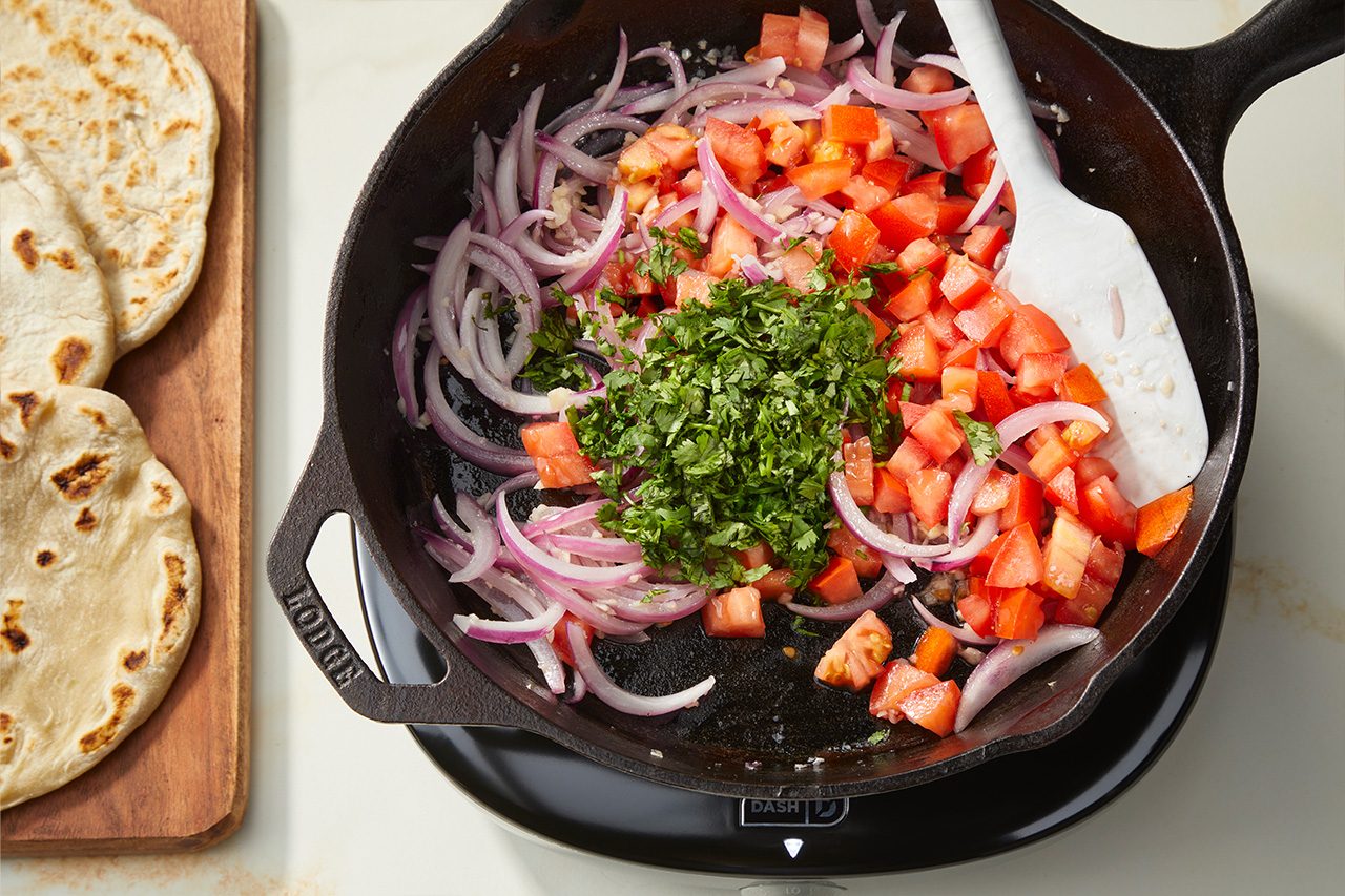 Overhead horizontal image of chopped red onions, diced tomatoes, and fresh cilantro inside a Lodge cast iron pan, situated on a black Dash portable burner. A white spatula rests among the ingredients, while a stack of flatbread is visible on a wooden cutting board to the left. | [Original] RMS 283003 East African Rolex; Rolled Eggs; Contest-Everything Eggs; How to; Step 3 Cook onion and garlic in pan; Show tomato and cilantro in pan and slightly softening; actionable shot is stirring; Horizontal