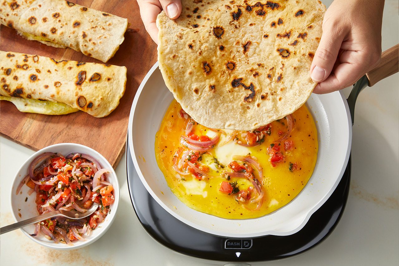 Overhead horizontal image of hands placing a grilled flatbread into a white pan, which contains a yellow egg mixture with chopped red onions and tomatoes. Beside the pan, a white bowl holds a vibrant salsa made of red onions, tomatoes, and herbs, and two finished flatbread wraps rest on a wooden cutting board. | [Original] RMS 283003 East African Rolex; Rolled Eggs; Contest-Everything Eggs; How to; Step 4 Non-stick pan (6") with eggs cooking in pan - actionable shot is showing topping with tomato mixture; edge in cooked chapati waiting to be put on top as well as some already rolled chapati/eggs.