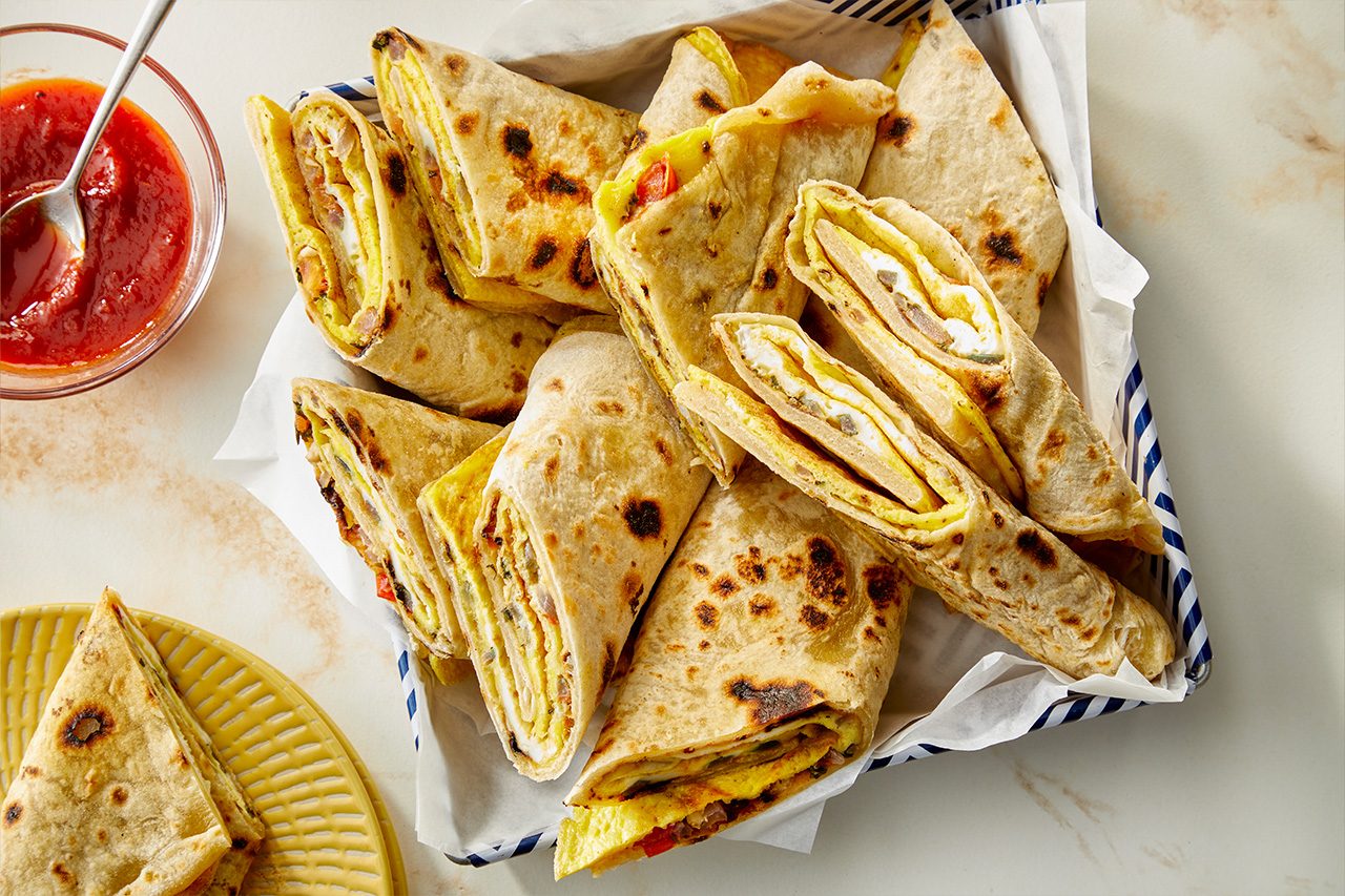 Overhead vertical image of several egg and vegetable-filled flatbread rolls nestled in a blue and white striped basket lined with white parchment paper. The rolls are cut in half, showcasing layers of cooked egg, diced red ingredients, and other savory fillings. A small glass bowl of red dipping sauce with a spoon and yellow plates are visible in the background. | [Original] RMS 283003 East African Rolex; Rolled Eggs; Contest-Everything Eggs; Vertical AP