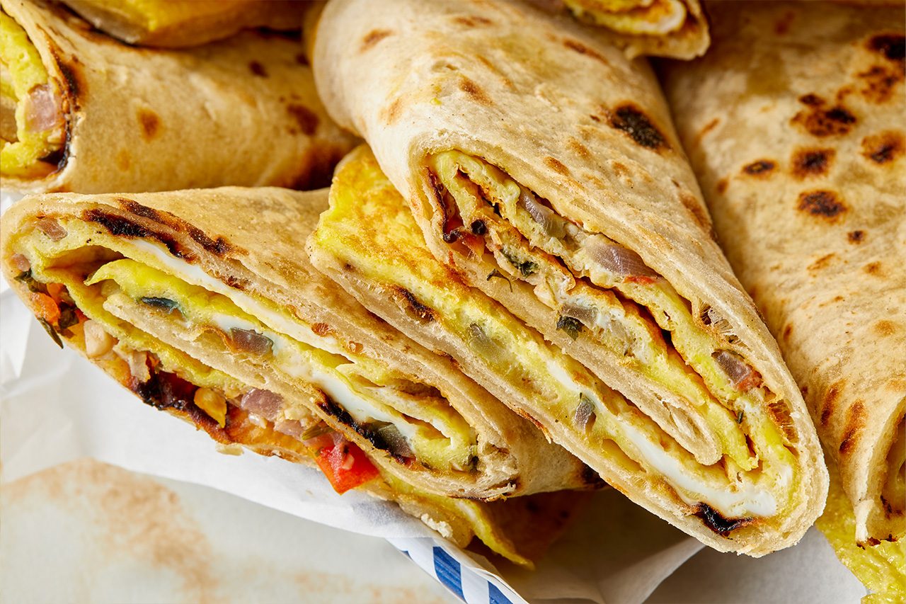 Overhead horizontal image of a basket filled with numerous egg-filled flatbread rolls, some cut to reveal their layered omelet interiors with visible onions and peppers. A small clear bowl of red sauce with a spoon is positioned to the left, and a yellow textured plate with an additional roll is visible in the bottom left corner. | [Original] RMS 283003 East African Rolex; Rolled Eggs; Contest-Everything Eggs; Horizontal AP; Pull