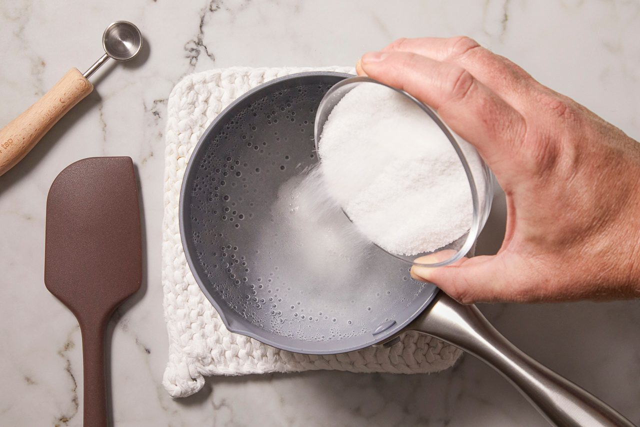 Overhead horizontal image of a hand pouring granulated sugar from a clear measuring cup into a gray saucepan containing water