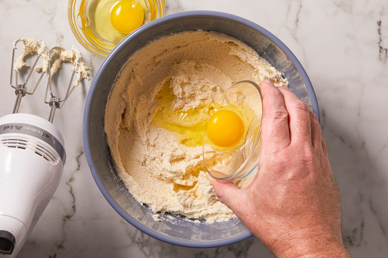 Overhead horizontal image of a hand pouring a raw egg from a small glass bowl into a larger blue mixing bowl filled with light-colored batter.