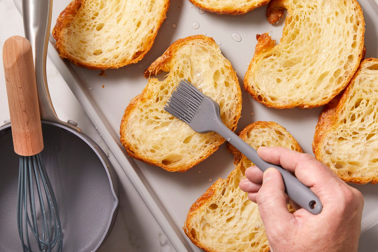 Overhead horizontal image of a hand brushing butter or oil onto multiple slices of golden-brown croissants arranged on a gray baking sheet.