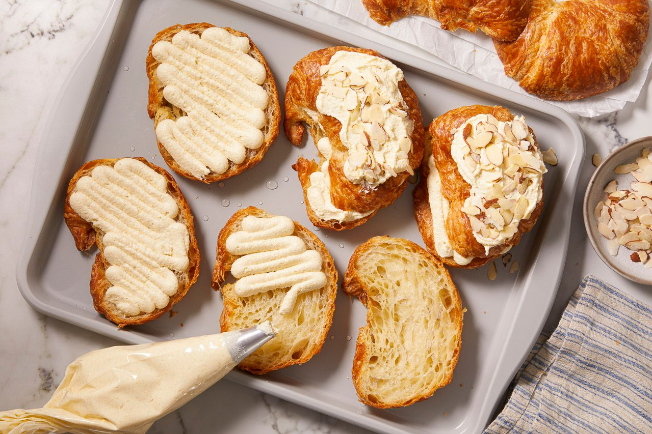Overhead horizontal image of several croissants filled with a piped creamy filling, some topped with sliced almonds, while another is in the process of being piped from a pastry bag.