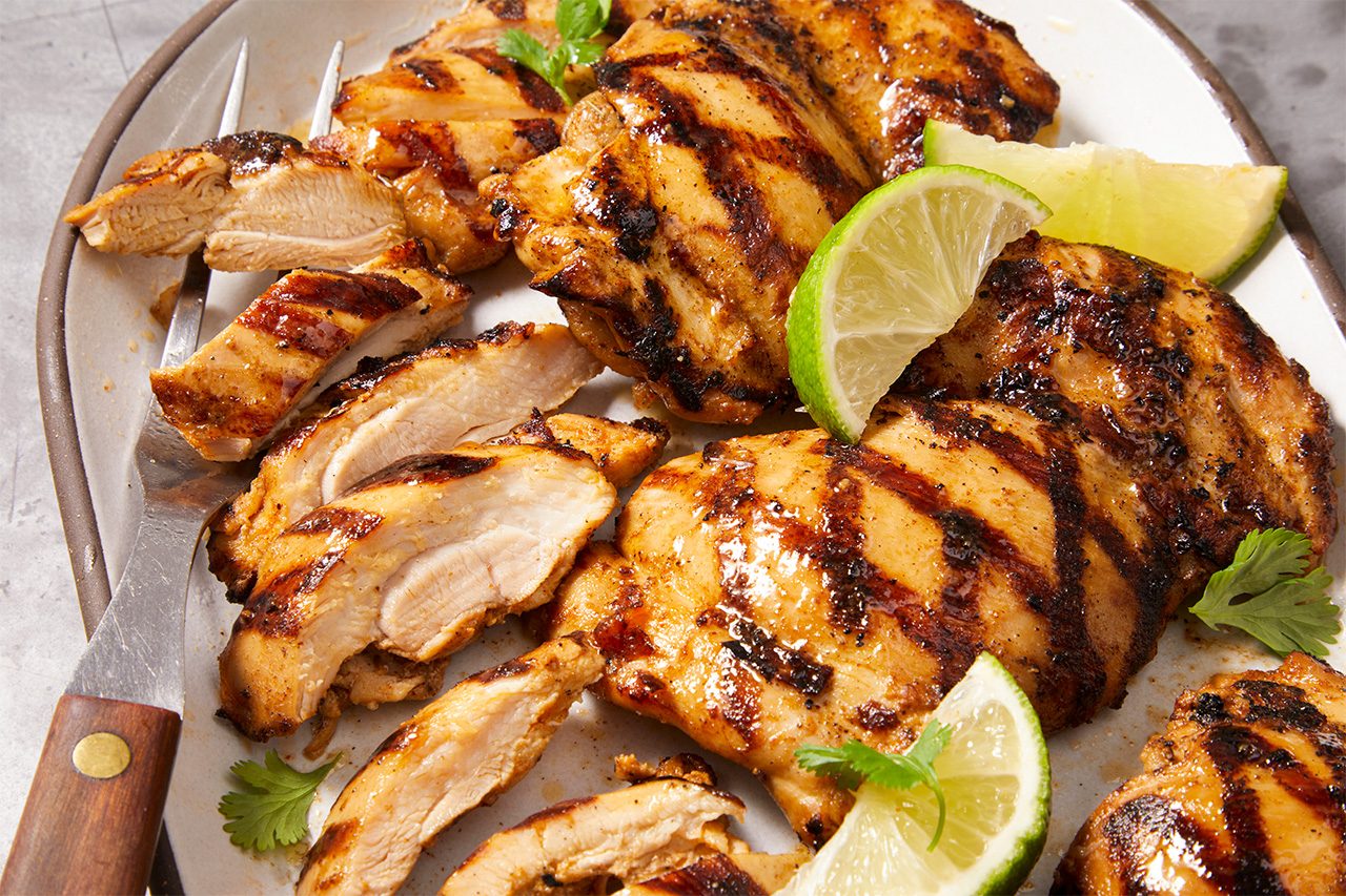 Eye level horizontal image of grilled chicken, some whole and some sliced, arranged on a plate. The chicken is garnished with fresh lime wedges and cilantro leaves, with a fork partially visible on the left.