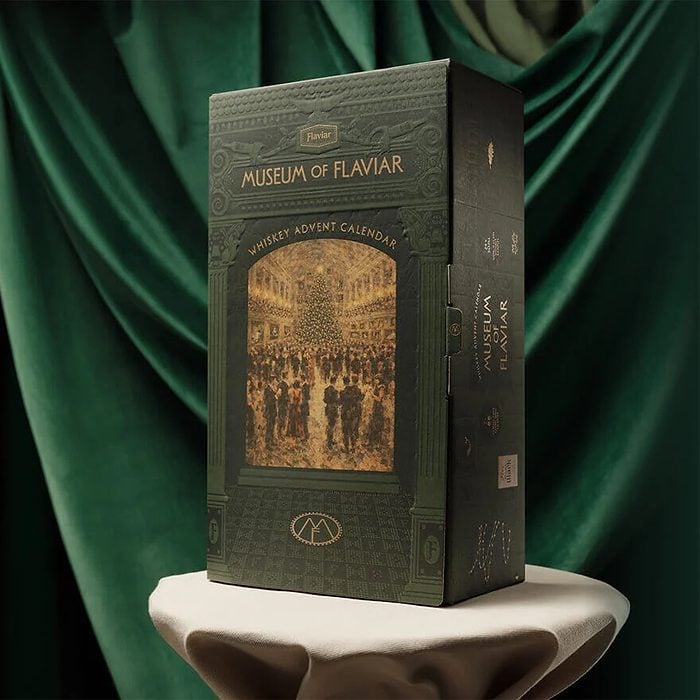 A green box labeled “Museum of Flaviar Whisky Advent Calendar” stands on a white pedestal, with green draped fabric in the background.