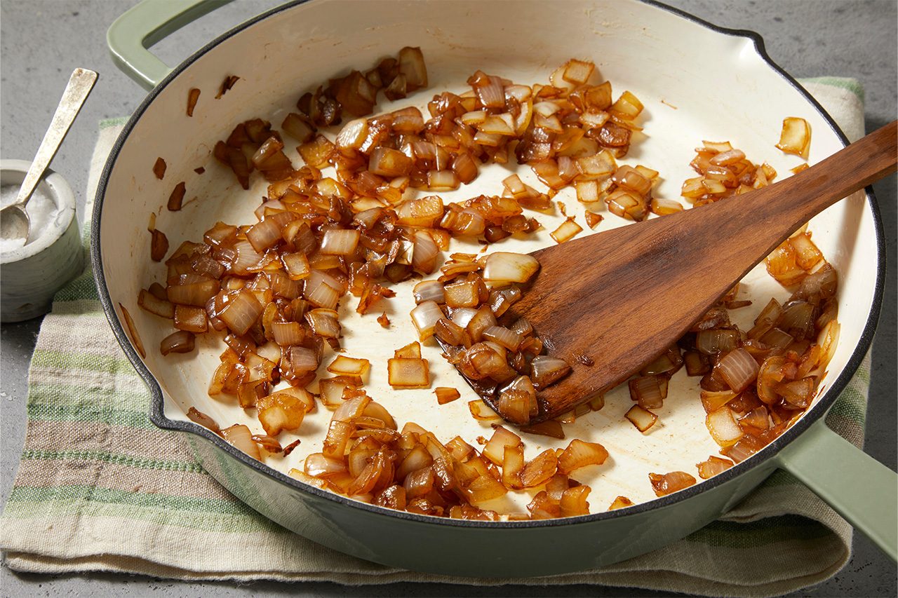 Overhead horizontal image of diced onions caramelizing in a light green enameled cast iron pan. A wooden spatula rests in the pan amidst the golden-brown, softened onions. A small bowl of salt and a striped linen napkin are visible in the background.