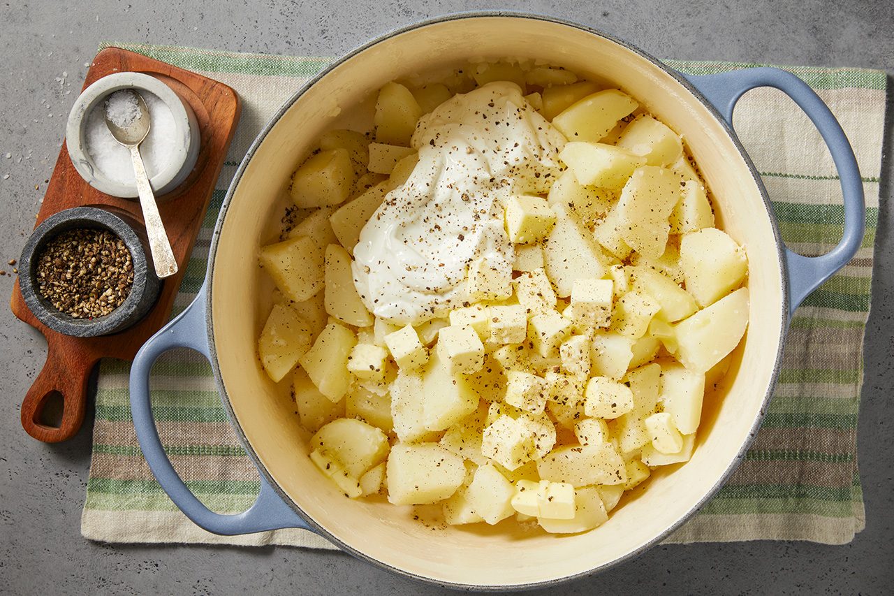 Overhead horizontal image of peeled, cubed potatoes in a light blue Dutch oven. A large dollop of sour cream or cream cheese, pats of butter, and freshly ground black pepper are added to the potatoes, ready for mashing. Small bowls of salt and pepper rest on a wooden board beside the pot.
