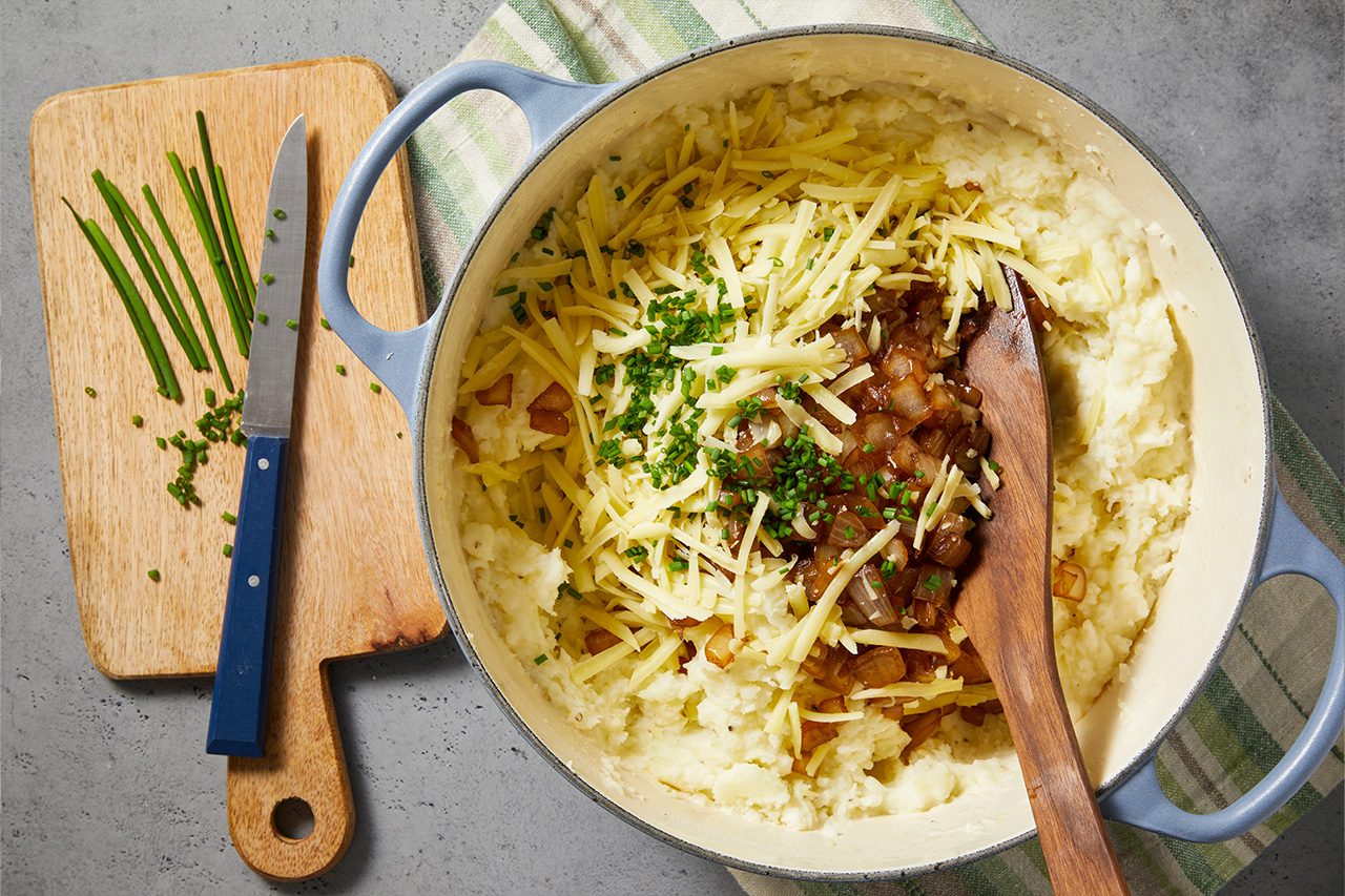 Overhead horizontal image of a light blue pot filled with creamy mashed potatoes, topped with shredded cheese, caramelized onions, and fresh chives. A wooden cutting board with a knife and more chives sits next to the pot, all resting on a striped towel and gray surface.