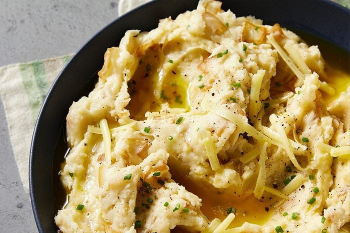 Eye Level horizontal image of a close-up of creamy, rustic mashed potatoes in a dark bowl, generously topped with shredded cheese, melted butter, chopped chives, and black pepper.