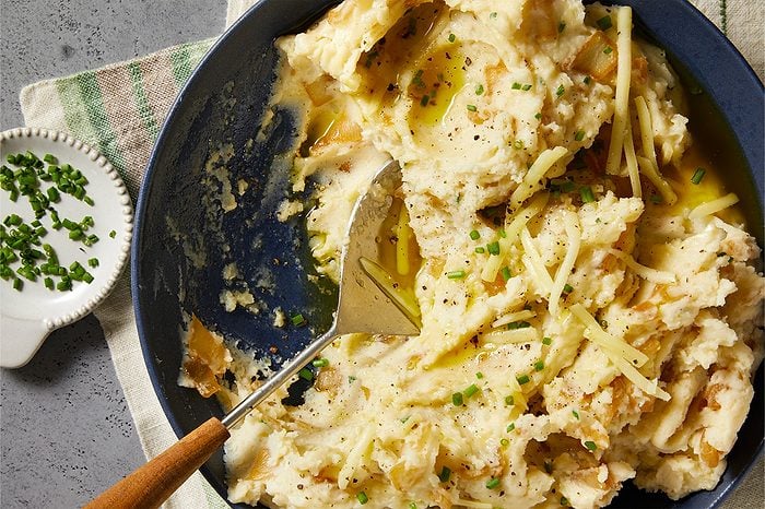Overhead horizontal image of creamy mashed potatoes served in a dark blue skillet. The potatoes are garnished with fresh chives, cracked black pepper, and shredded cheese, with olive oil drizzled over the top. A serving spoon rests in the potatoes, and a small dish of additional chopped chives sits nearby on a gray countertop with a striped napkin.