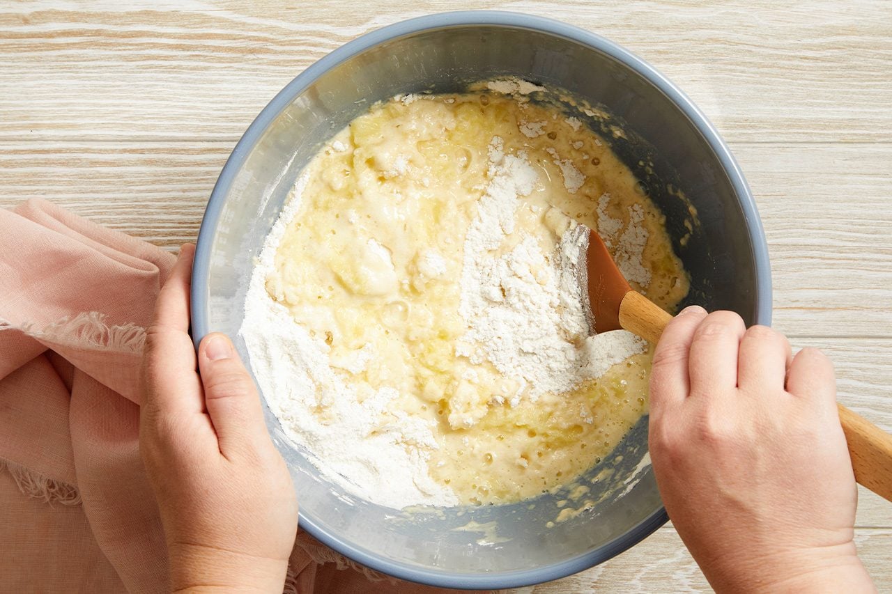 Overhead horizontal image of hands mixing ingredients in a gray bowl on a light wooden surface. A person's hands are stirring a wet, lumpy mixture with a brown-headed wooden spatula, incorporating a pile of white flour, indicating the preparation of a baked good batter.