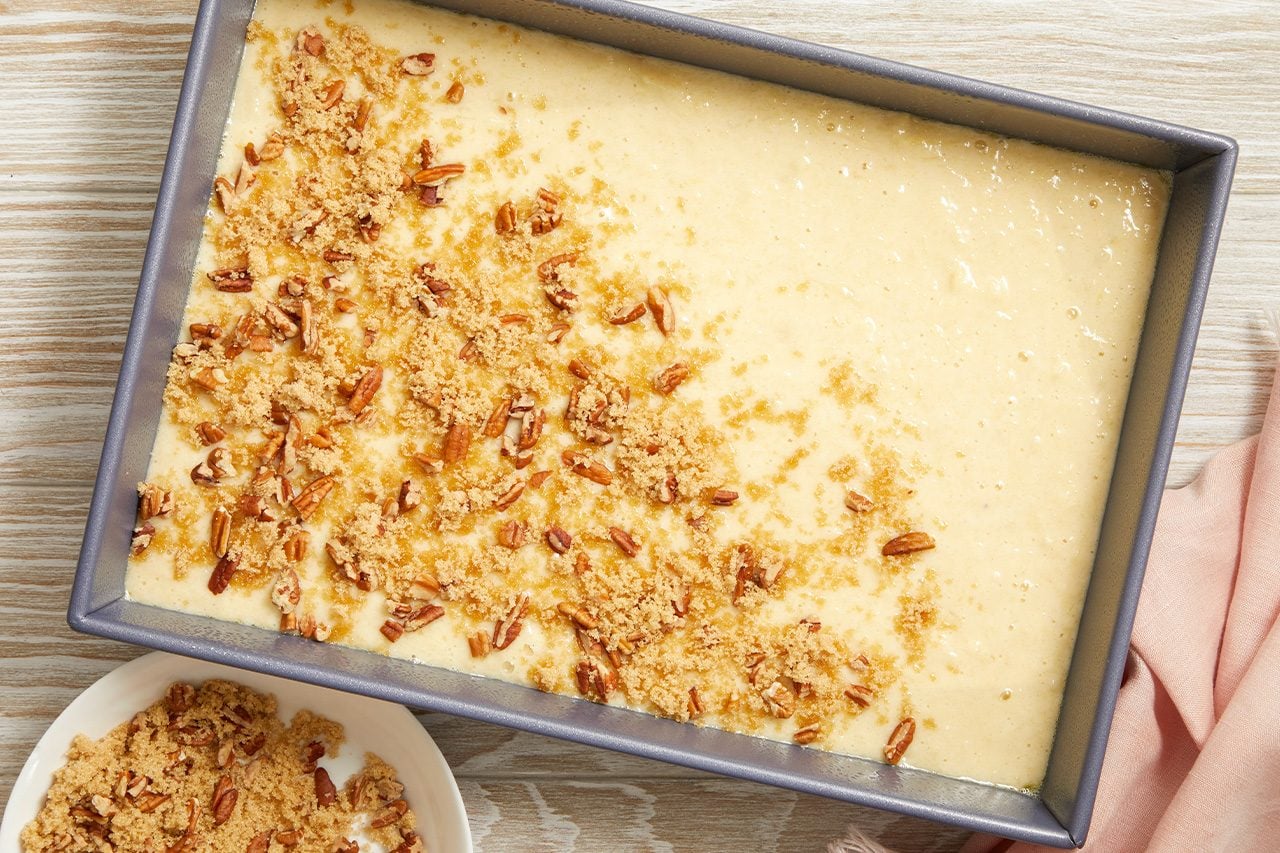 Overhead horizontal image of a rectangular baking pan filled with light-colored batter. One half of the batter is covered with a topping of brown sugar and chopped pecans, while a white bowl in the foreground holds more of the topping mixture.