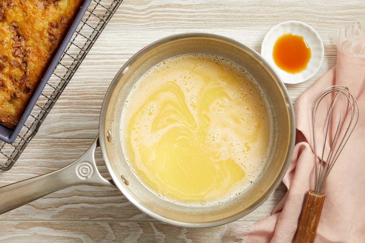 Overhead horizontal image of a saucepan containing a bubbling melted butter mixture on a light wooden surface. A small white dish with amber liquid, a whisk on a pink cloth, and a baked coffee cake in a purple dish on a cooling rack are also visible.