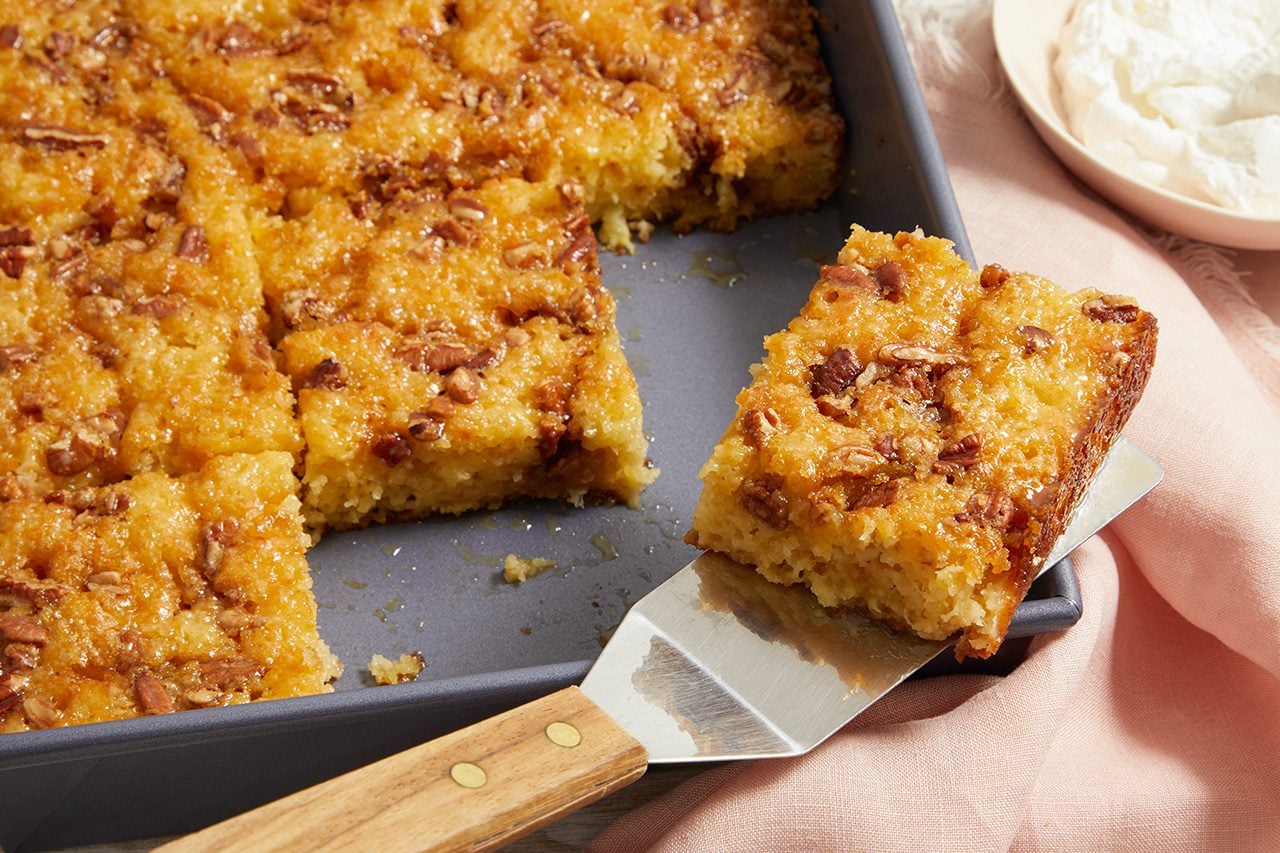 Overhead horizontal image of a delicious-looking sheet cake. A slice of golden brown cake, topped with chopped pecans and a sweet glaze, is being lifted from a gray baking pan with a metal spatula. A bowl of whipped cream is visible in the soft-focused background, ready to accompany the dessert.